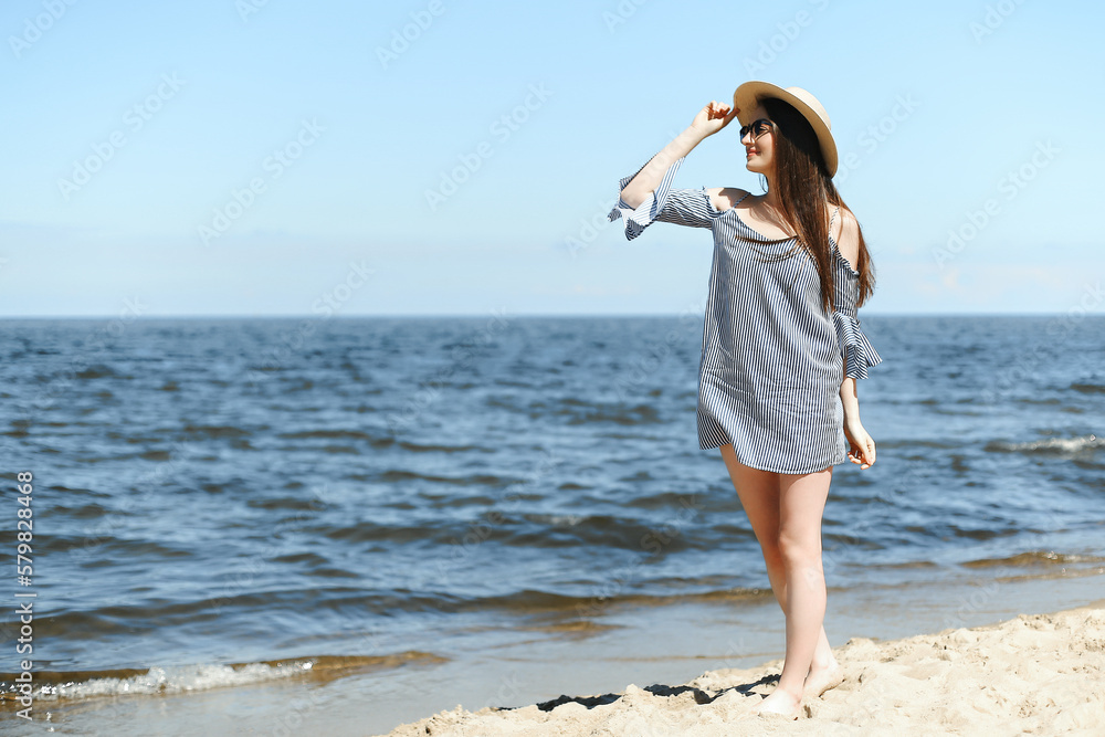 Happy young brunette woman standing on the ocean beach while smiling, and wearing fashion hat, sunglasses. The enjoying vacation concept. Happy young brunette woman standing on the ocean beach while smiling, and wearing fashion hat, sunglasses. The enjoying vacation concept.