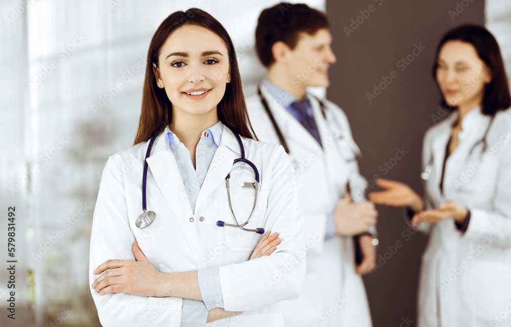 Smiling intelligent woman-doctor is standing with arms crossed in a clinic, together with her colleagues at the background. Portrait of physicians at work. Perfect medical service in a hospital Smiling intelligent woman-doctor is standing with arms crossed in a clinic, together with her colleagues at the background. Portrait of physicians at work. Perfect medical service in a hospital