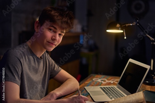 Teenage student learning at home with books and laptop computer. Student in online school and education, sitting at desk late night