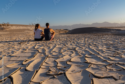 Loving couple sitting on dry cracked clay crust at Mesquite Flat Sand Dunes in Death Valley National Park, California, USA. Looking at Mojave desert in summer with Amargosa Mountain Range in the back photo