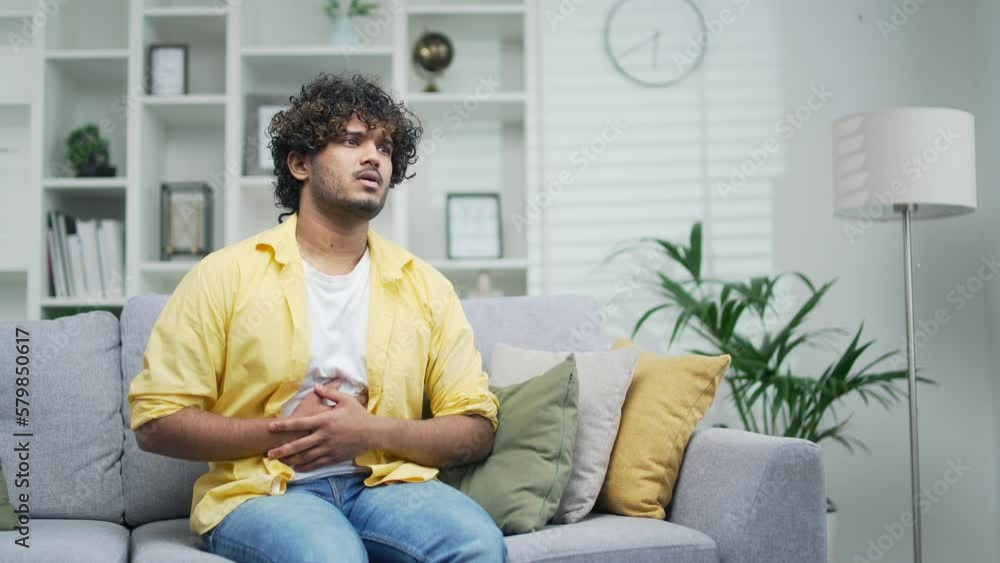 A handsome young man suffers from abdominal pain sudden ache poisoning diarrhea indigestion holding his stomach while sitting at home indoor People gastritis problem constipation touching belly A handsome young man suffers from abdominal pain sudden ache poisoning diarrhea indigestion holding his stomach while sitting at home indoor People gastritis problem constipation touching belly