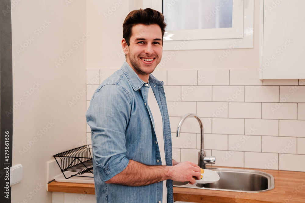Young caucasian male washing the dishes in a home kitchen. Handsome smiling man doing housework cleaning and looking at camera.. Housekeeping concept. High quality photo