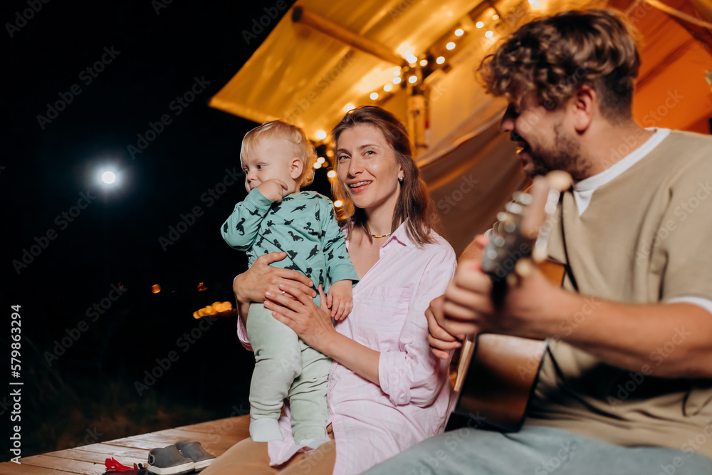 Happy family relaxing and spend time together in glamping on summer evening and playing guitar near cozy bonfire. Luxury camping tent for outdoor recreation and recreation. Lifestyle concept