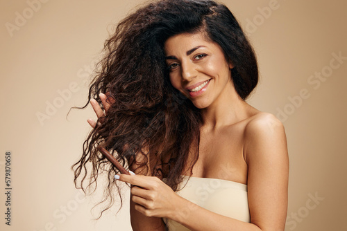HAIR MORNING ROUTINE CONCEPT. Enjoyed happy awesome curly Latin lady with hairbrush combing curls smiling posing isolated on pastel beige background, look at camera. Copy space, good cosmetic offer