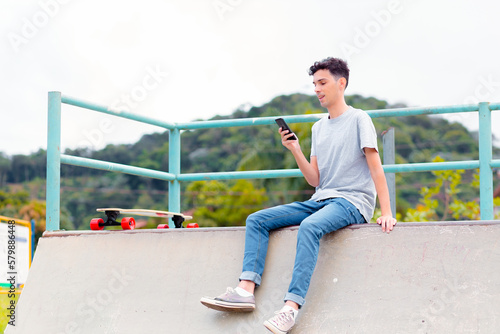 Happy man sitting on top of a skate ramp, with a longboard on his side, while using his cell phone or smartphone to chat online or access social networks.