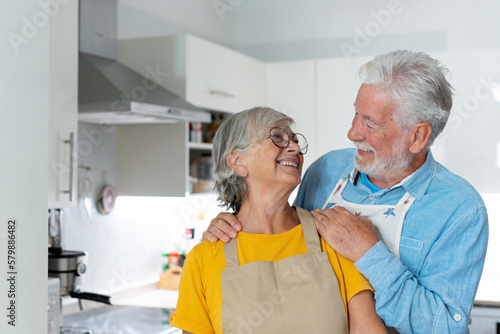 Head shot portrait smiling mature married couple looking at camera  standing in kitchen cooking at home  happy senior man wearing glasses hugging wife  grandparents posing for photo.