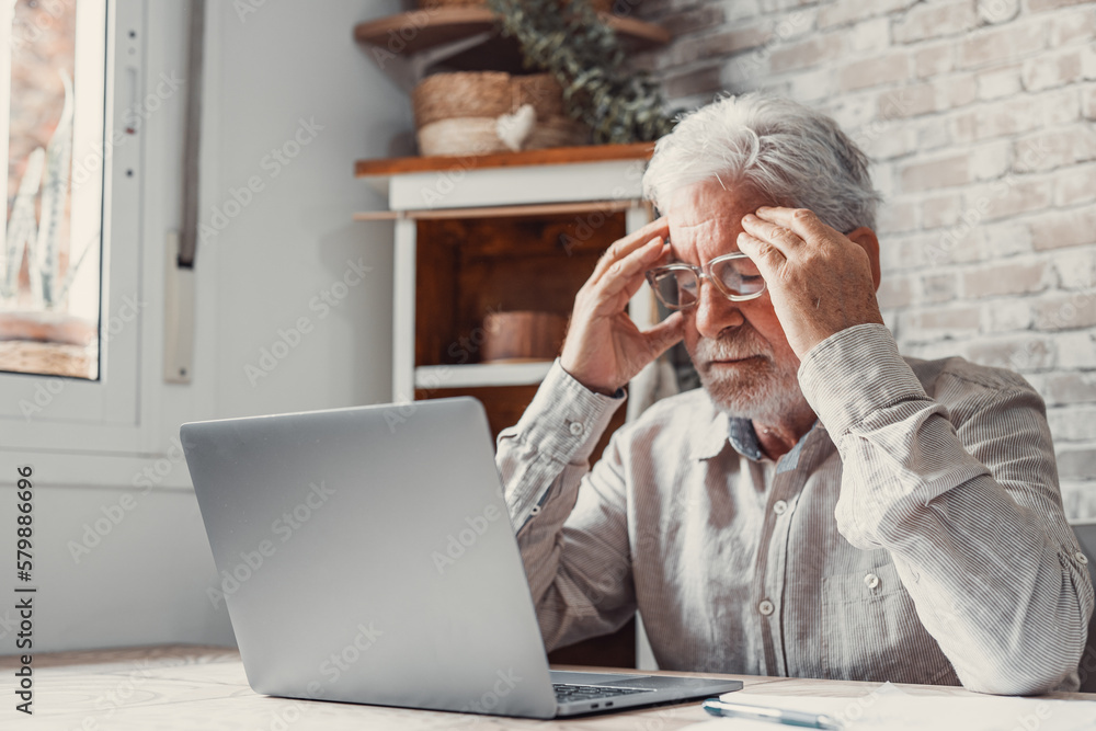 Tired mature 70s man working at laptop from home, taking off glasses