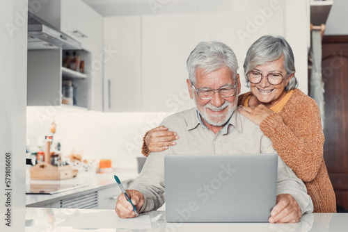 Old age and modern tech. Retired family couple sit at kitchen table use laptop watch photo video read latest news online. Aged husband share discuss interesting information on website with senior wife