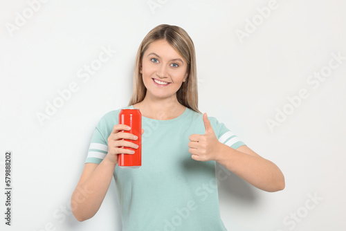 Beautiful happy woman holding red beverage can and showing thumbs up on light background