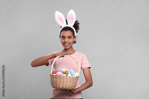 Happy African American woman in bunny ears headband holding wicker basket with Easter eggs on gray background