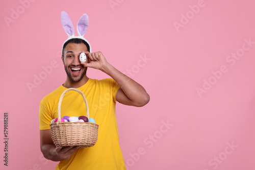 Happy African American man in bunny ears headband covering eye with Easter egg on pink background, space for text