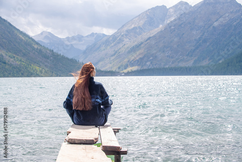Young tourist woman is sitting on pier near mountain lake and looking at a beautiful landscape. Back view