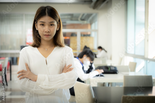 The beautiful asian female teacher is standing with arms crossed and looking at the camera with a serious face in the library room. © Digital Art Studio