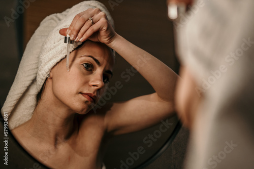 A beautiful woman is using anti aging serum treatment on her face and looking at herself in the mirror in the bathroom.