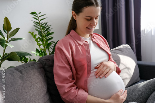 Happy maternity concept. Smiling young single pregnant woman sitting on cozy couch in light room touch big belly enjoy late stages of pregnancy. Caring mother strokes pregnant tummy with two hands