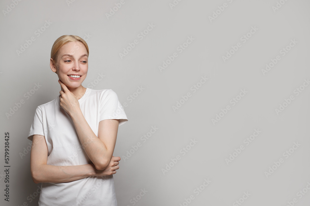 Attractive blonde female model in white t-shirt looking aside and smiling against grey studio wall banner background Attractive blonde female model in white t-shirt looking aside and smiling against grey studio wall banner background