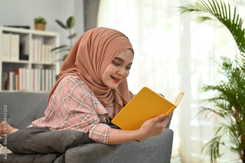Happy young Muslim woman in hijab and casual clothes laying on couch and reading a book. People and leisure activity concept Happy young Muslim woman in hijab and casual clothes laying on couch and reading a book. People and leisure activity concept