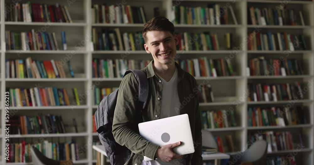 Cheerful handsome college student guy standing in library, holding laptop, backpack, looking at camera, turning at window away, smiling, laughing with bookshelves in background Cheerful handsome college student guy standing in library, holding laptop, backpack, looking at camera, turning at window away, smiling, laughing with bookshelves in background