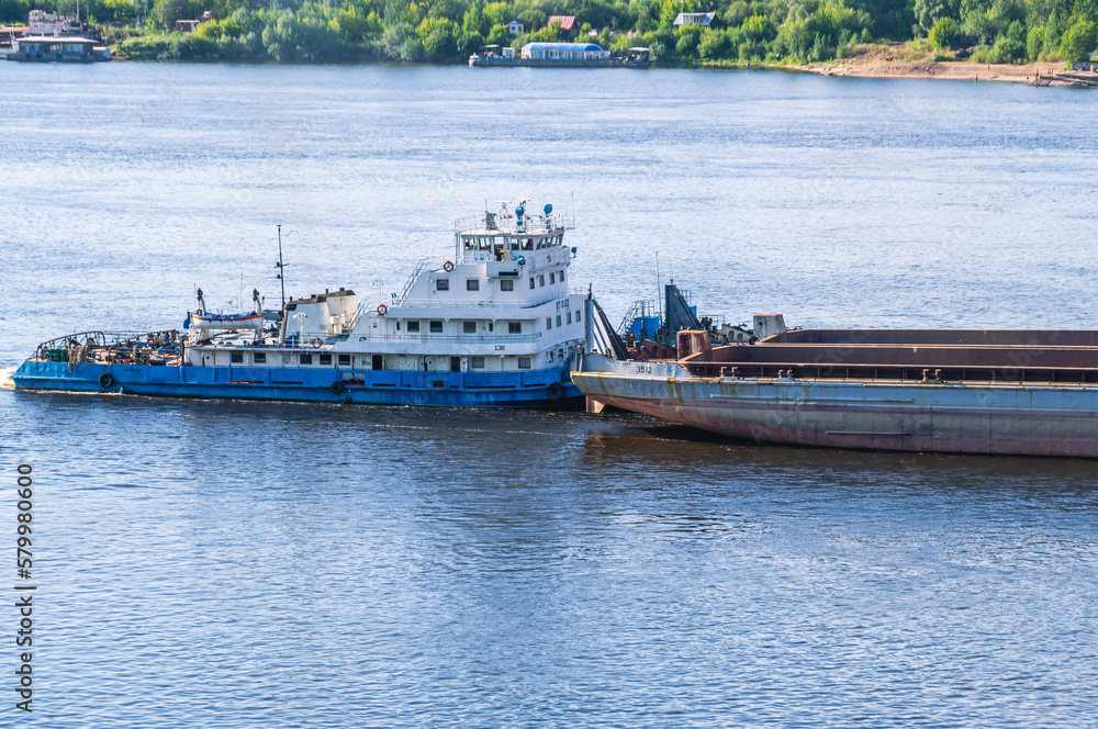 A tugboat pushes a large barge with cargo along a wide river. In summer, the tugboat goes down the river. Cargo transportation by river. Navigation of vessels on the river. A tugboat pushes a large barge with cargo along a wide river. In summer, the tugboat goes down the river. Cargo transportation by river. Navigation of vessels on the river.