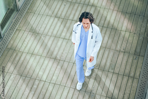 病院の廊下を歩くスクラブ白衣を着た若い男性医師 © HML