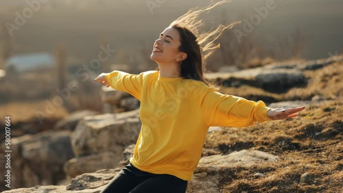 A young woman in a yellow sweater sits on the edge of a rock and raises her arms to the sides imitating flight, happy and cheerful admiring nature, wind blowing her hair in the sunlight, traveling