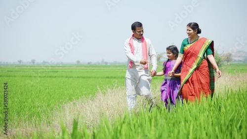 Happy village couple with kid walking by talking each other near paddy field - concept of sustainable lifestyle, family bonding and rural India.