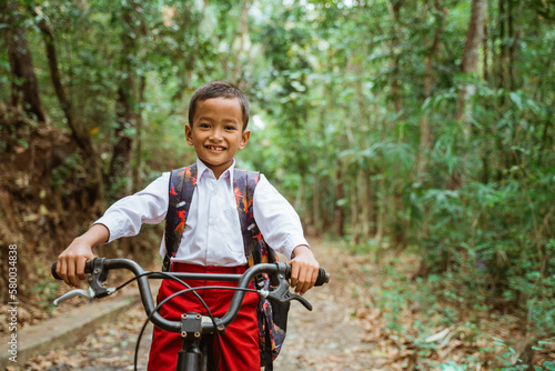 a male elementary student in uniform riding his bike through the country road and smiling with a lot of tree at the background