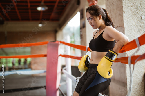 a female boxer with yellow boxing gloves leaning on the ropes inside the box ring