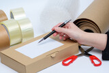 Karton z przesyłką i etykietą na tle akcesoriów do pakowania paczek takich jak folia bąbelkowa, taśma, nożyczki i dłoń z długopisem w sklepie internetowym 