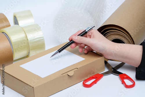 Karton z przesyłką i etykietą na tle akcesoriów do pakowania paczek takich jak folia bąbelkowa, taśma, nożyczki  i dłoń z długopisem w sklepie internetowym  photo