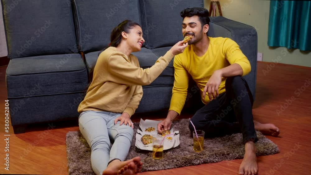 Happy young couple feeding each other with pizza while sitting on floor at home - concept of dating, affection and love relationship. Happy young couple feeding each other with pizza while sitting on floor at home - concept of dating, affection and love relationship.