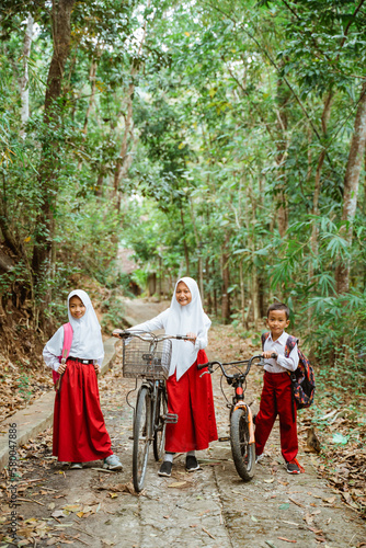 three elementary students in uniform standing together at the country road bring their bike with a lot of tree around them