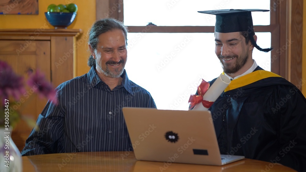 Φωτογραφία Proud happy father and happy son sitting at dining room table doing online graduation ceremony concept in costume and hugging