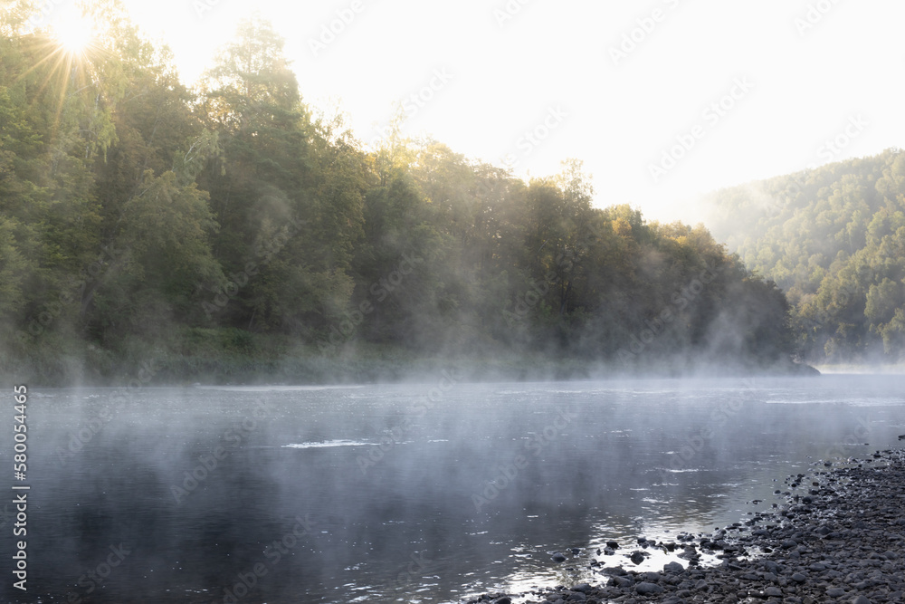 Tranquil bright early morning landscape with mist on lake - soft cold haze on water, lush green forest in golden sunbeams, pebble shore. Breathtaking beautiful sunrise in wild nature for recreation. Tranquil bright early morning landscape with mist on lake - soft cold haze on water, lush green forest in golden sunbeams, pebble shore. Breathtaking beautiful sunrise in wild nature for recreation.