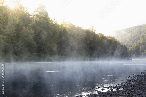 Tranquil bright early morning landscape with mist on lake - soft cold haze on water, lush green forest in golden sunbeams, pebble shore. Breathtaking beautiful sunrise in wild nature for recreation.