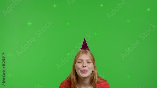 Attractive red-haired white girl in a party hat celebrates her birthday against the backdrop of a green screen. Happy young woman in jeans and knitted jumper blowing out candles on virtual cake photo