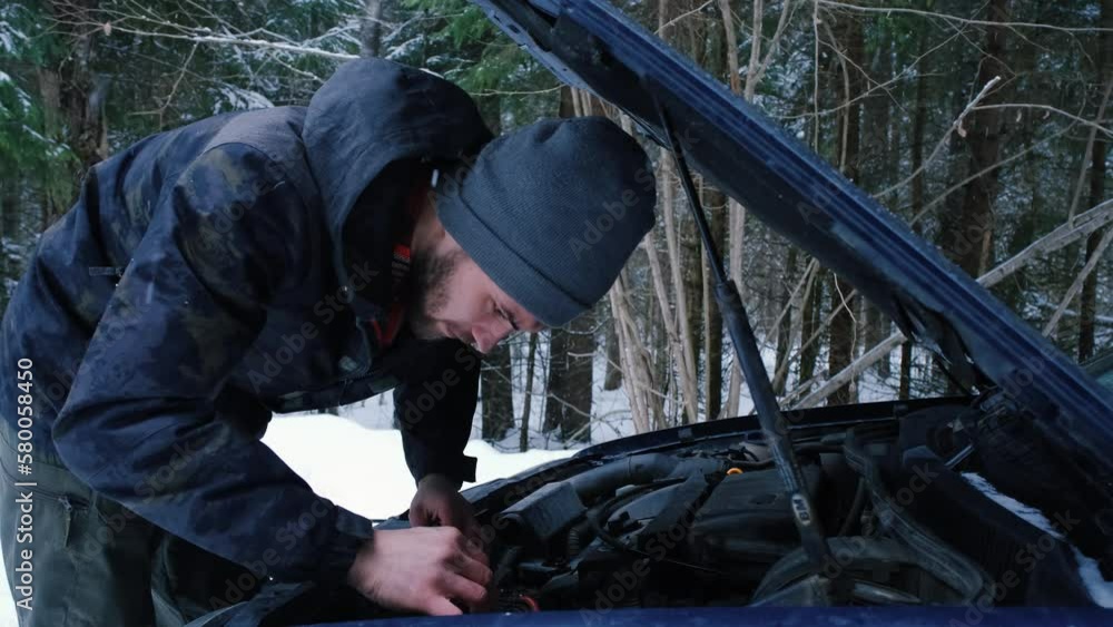 Young Caucasian man has opened hood of car and looking for cause of breakdown while standing in snowy forest in winter. Guy driver checks his transport before trip. Concept problem of old used cars. Young Caucasian man has opened hood of car and looking for cause of breakdown while standing in snowy forest in winter. Guy driver checks his transport before trip. Concept problem of old used cars.