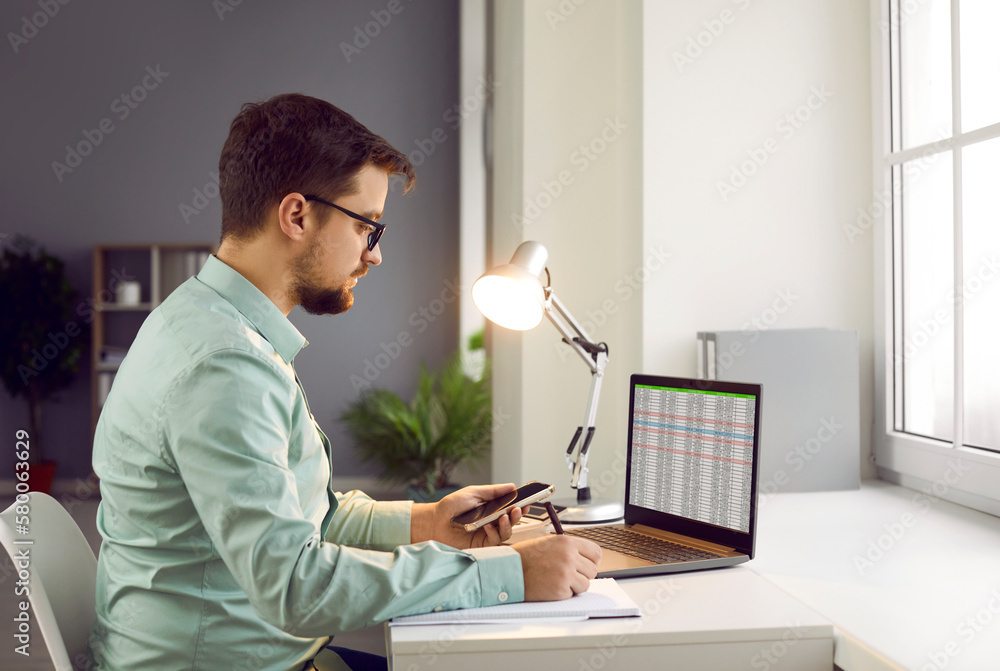 Portrait of serious businessman working on documents in office. Man in eyeglasses sitting at desk in front of computer, holding smartphone and making notes on paper documents