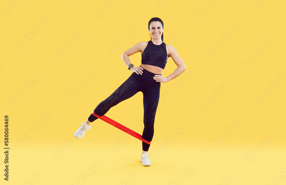 Fitness woman standing and stretching elastic band with legs isolated on colored background. Full length woman with sporty body dressed in black leggings and sport top training on yellow background.