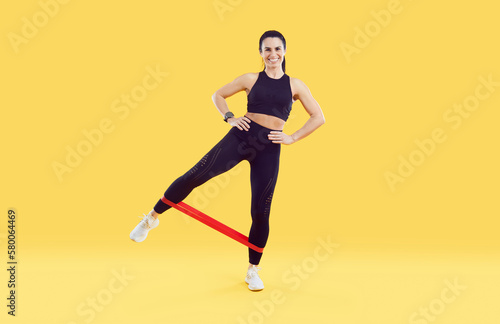 Fitness woman standing and stretching elastic band with legs isolated on colored background. Full length woman with sporty body dressed in black leggings and sport top training on yellow background.