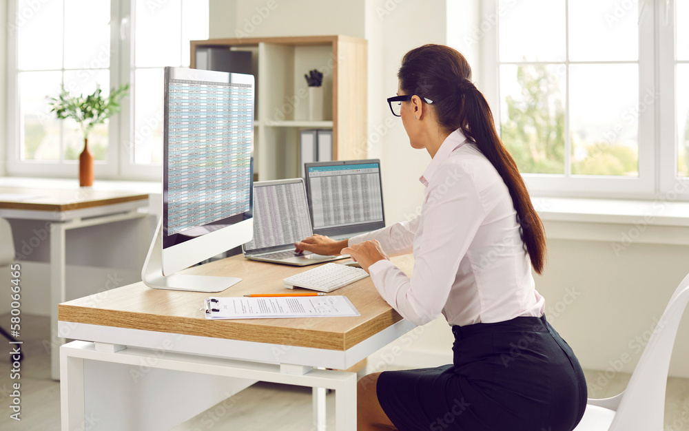 Side view portrait of a brunette attractive young business woman accountant sitting at the desk and working on a laptop with tables and charts at office and looking to the monitor screen.