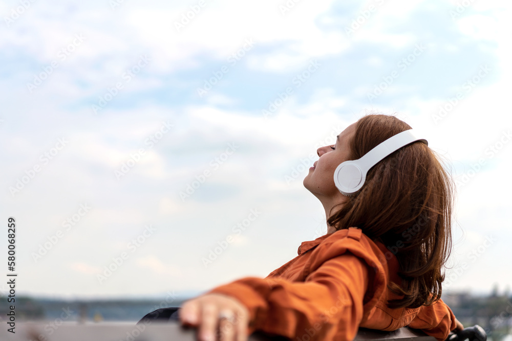 Woman sitting on bench outdoors listening music using wireless headphones.