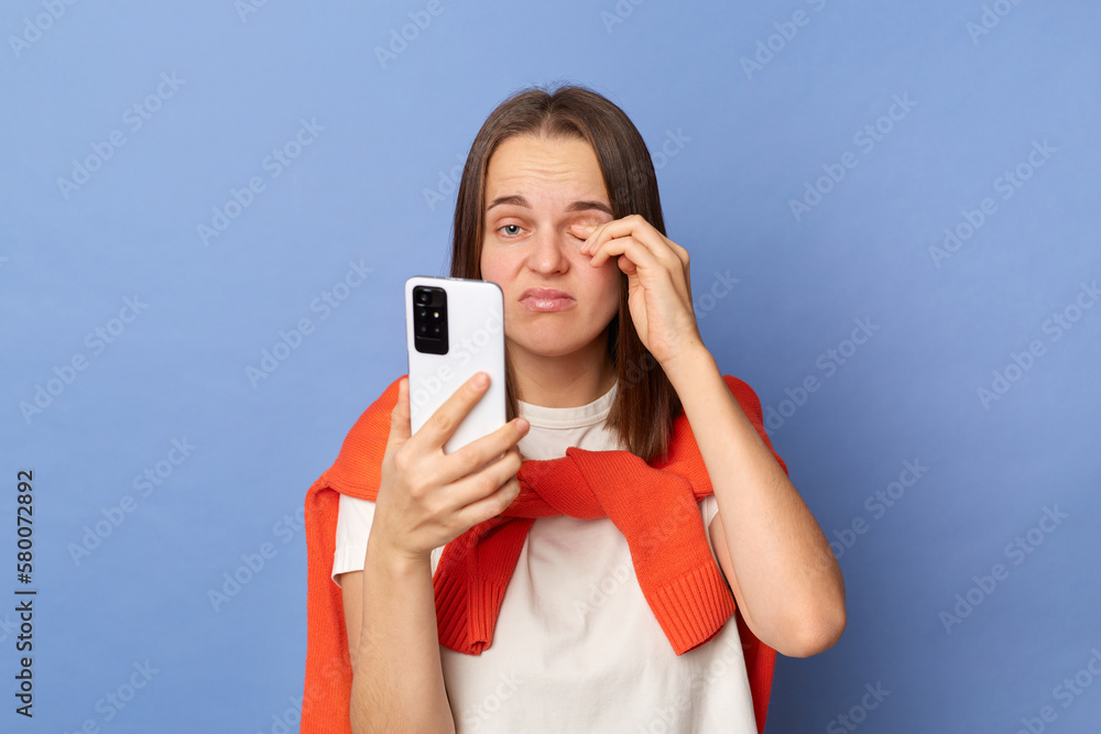 Indoor shot of tired sad woman wearing white t-shirt and jumper over shoulders using mobile phone, feels unwell and eyes hurt, rubbing her eyes, frowning face.