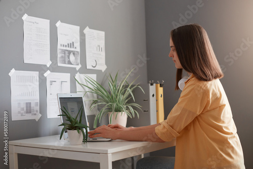 Slika na platnu Image of serious woman freelancer wearing yellow shirt having brown hair typing on notebook in office, having concentrated facile expression, doing report or working on project