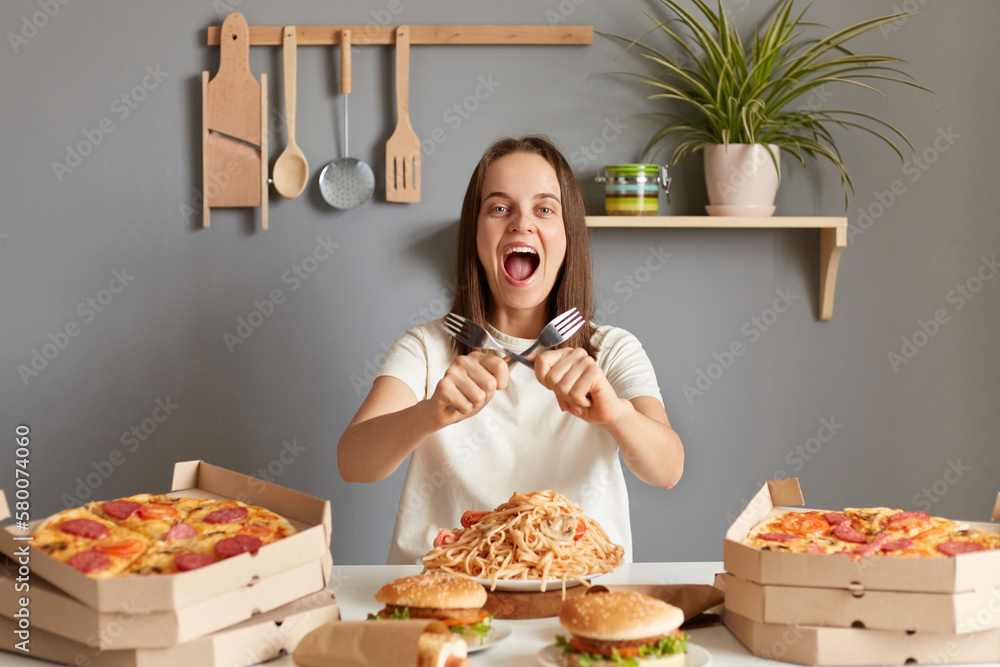 Portrait of amazed overjoyed woman with brown hair wearing white casual T-shirt sitting at table in kitchen, holding to forks, crossed hands, yelling with crazy expression, being extremely hungry. Portrait of amazed overjoyed woman with brown hair wearing white casual T-shirt sitting at table in kitchen, holding to forks, crossed hands, yelling with crazy expression, being extremely hungry.