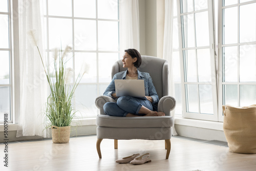 Thoughtful pretty middle aged lady relaxing with laptop in armchair, looking at window away, dreaming, enjoying leisure, calmness. Freelance business woman thinking on project, using computer at home