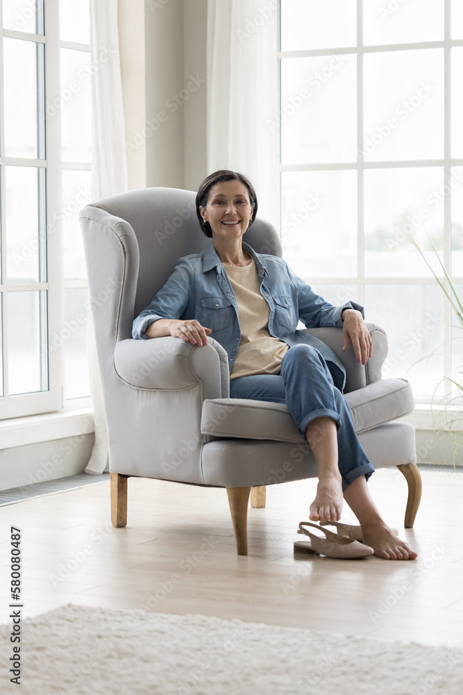 Happy pretty mature woman in casual resting in armchair home portrait. Pretty black haired senior lady looking at camera, smiling, enjoying leisure at large window in modern house. Full length
