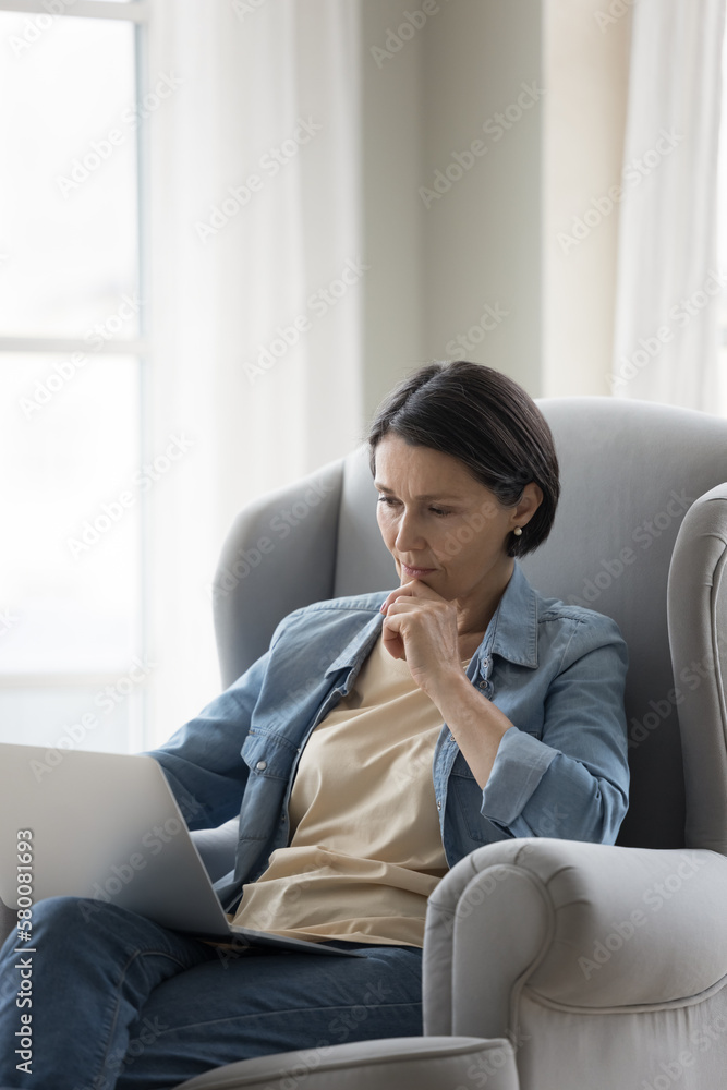 Serious pensive mature freelance woman working at laptop, sitting in comfortable armchair, looking at display, screen, touching chin, thinking over online content, Vertical shot