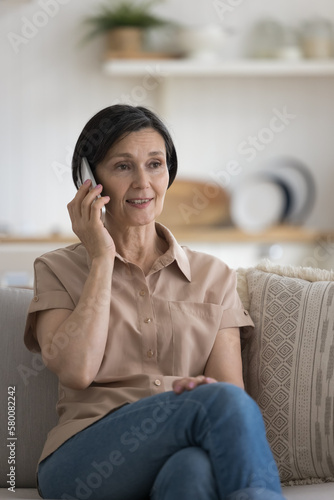 Pensive positive senior lady talking on mobile phone, making call, enjoying conversation, leisure on home sofa. Mature customer woman speaking to client support service on cell