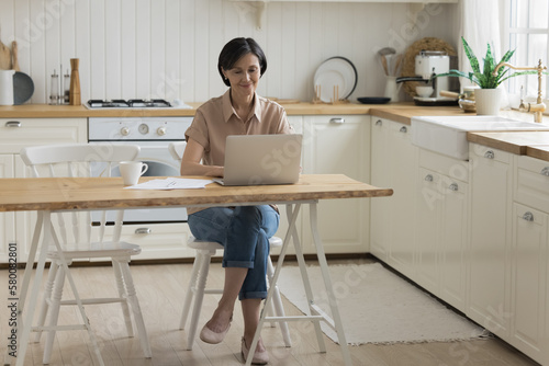 Positive focused mature freelance professional woman working at laptop from home, sitting at table in kitchen interior, typing, smiling. Senior homeowner using online service for domestic payment